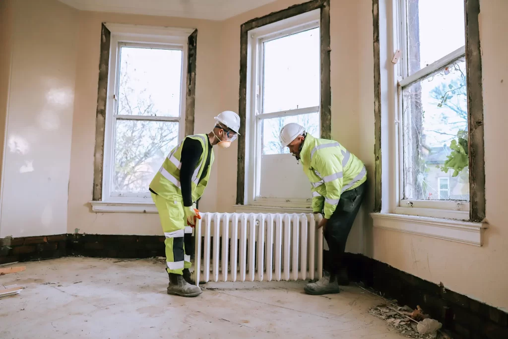 Two men wearing safety gear carry out an old iron radiator from a house.